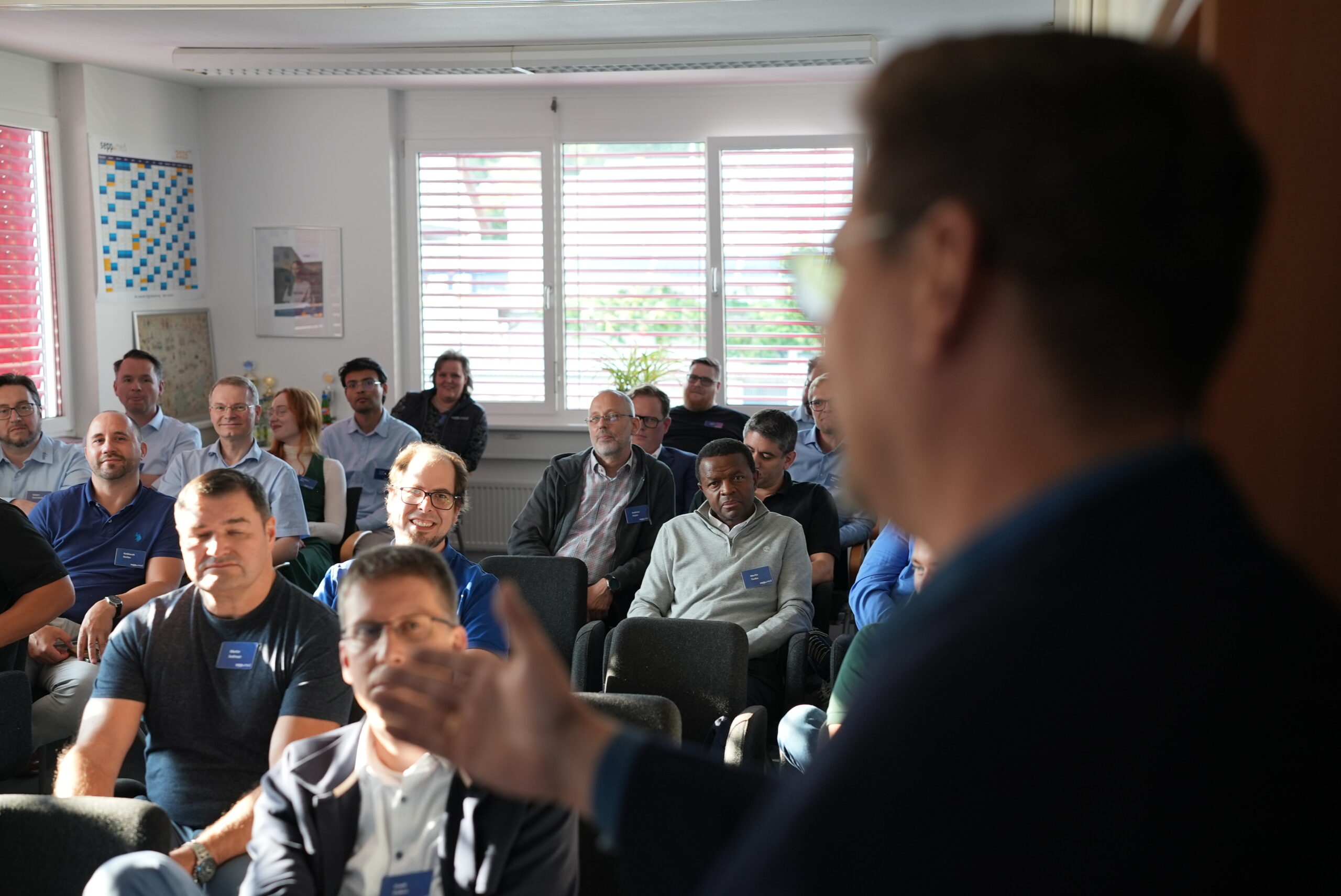 Eine Gruppe von Menschen sitzt in Reihen und hört aufmerksam einem Redner zu, der in einem hellen Raum mit großen Fenstern und einem Whiteboard an der Wand steht. Der Redner ist teilweise im Vordergrund zu sehen.